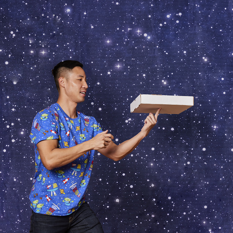 Man wearing the blue Aliens Unisex Tee and balancing a pizza box on his finger against a starry background